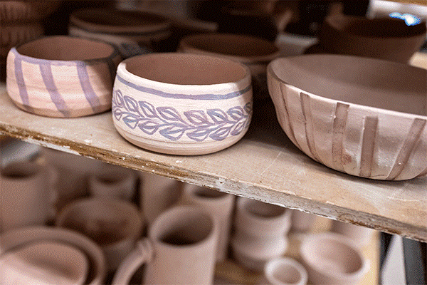 A brown shelf is visible with rows of unglazed pottery. The pottery pieces are shaped into bowls. The bowl on the left has five stripes visible from top to bottom. The bowl in the center has blue floral designs. The bowl on the right has three blue stripes slanted from top to bottom. Below this shelf is another shelf visible with clay pottery pieces.