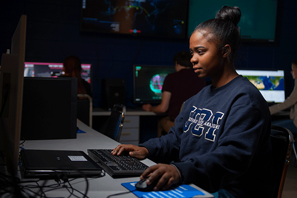 Female student of color with black hair in a bun and wearing a navy Indiana State University sweatshirt works on computer in a dark computer lab. Illuminated computer terminals and large-screen monitors are visible behind her.